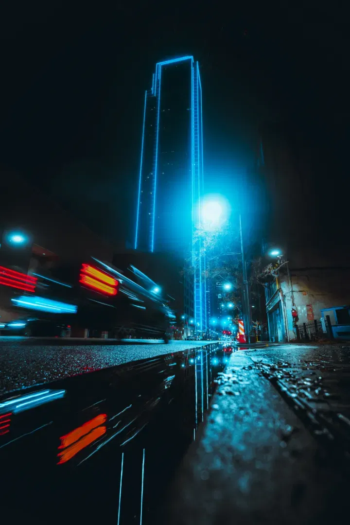 A dynamic shot from street level showing a blurred train passing in front of a neon-glowing high-rise at night.