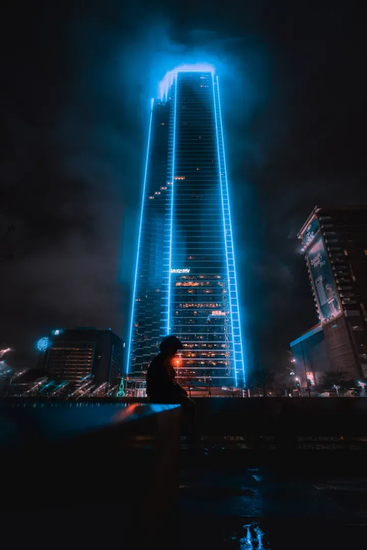 Low angle view of a blue neon-outlined skyscraper at night, with a silhouette of a person sitting on a ledge.
