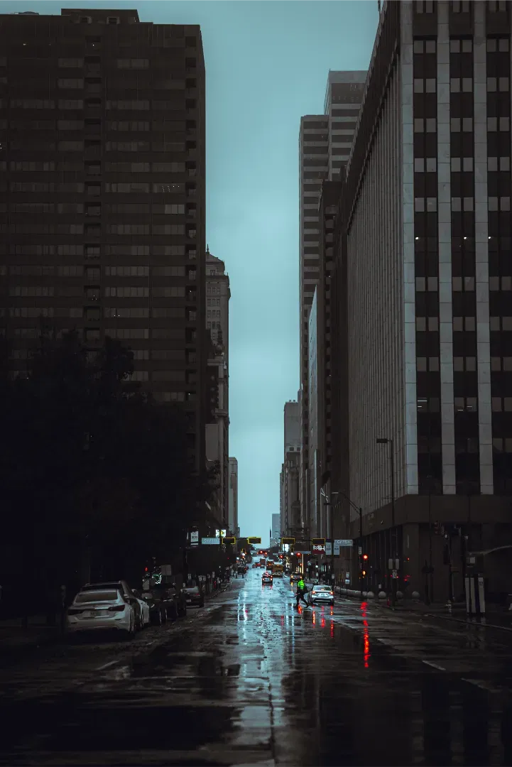 Looking down a wet city street flanked by tall skyscrapers under an overcast sky.