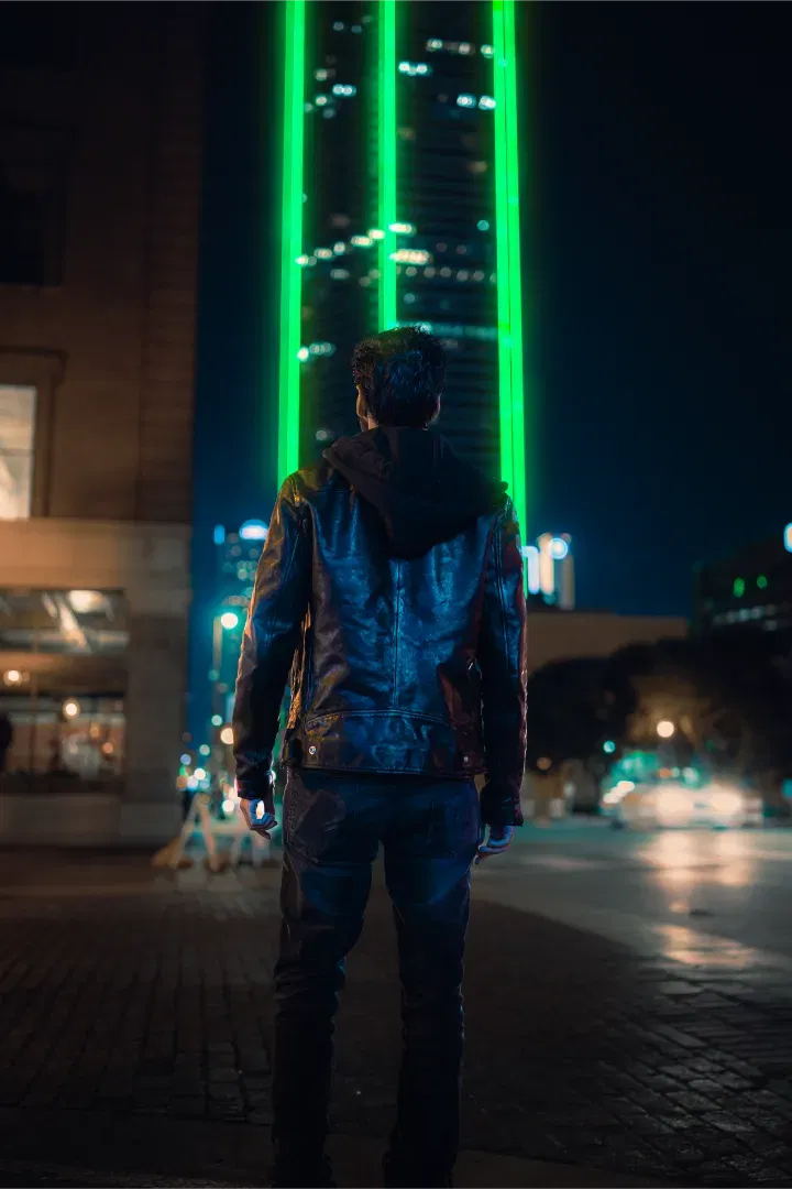 A man in a leather jacket standing with his back to the camera, looking at the green neon-lit Bank of America Plaza.