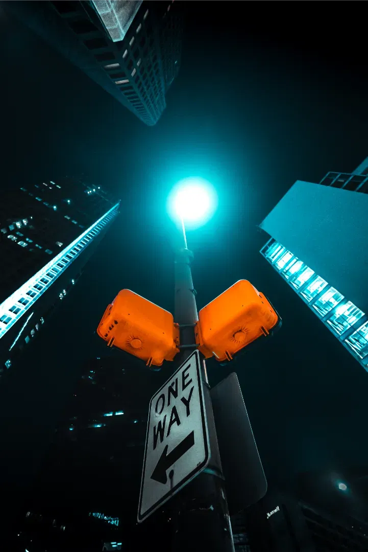 Low angle upward shot of a ONE WAY sign and traffic signal box under a bright streetlamp at night.