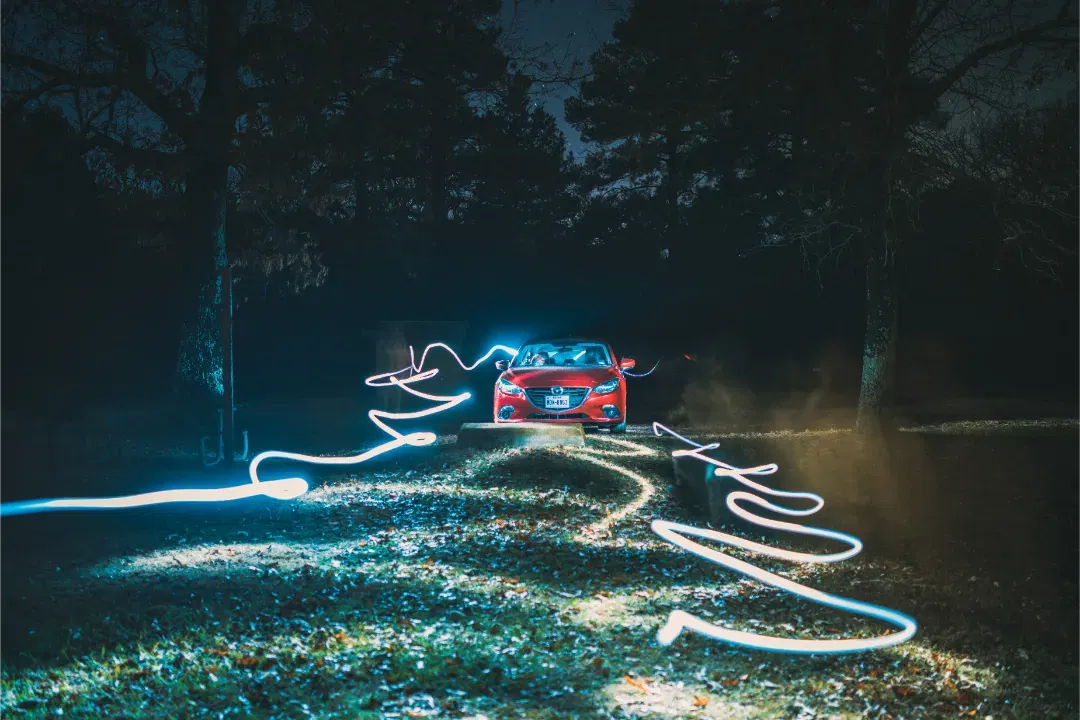 A red car parked in a dark wooded clearing at night, with swirling white long exposure light painting patterns.