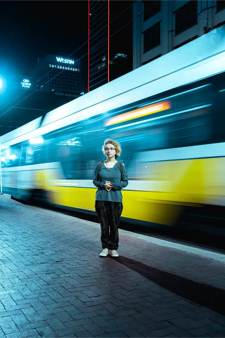 A woman standing on a transit platform as a fast-moving, yellow and blue light-rail train blurs behind her.