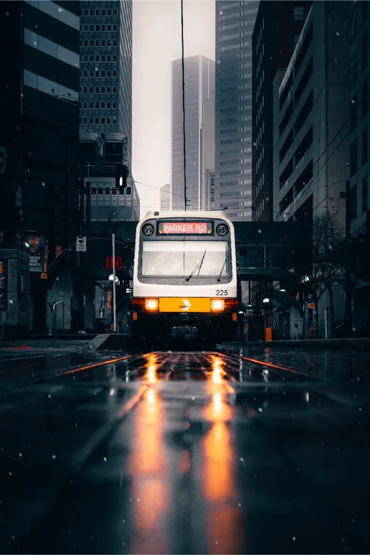 A light rail train approaching a street-level platform at night, with warm headlights reflecting in wet asphalt.