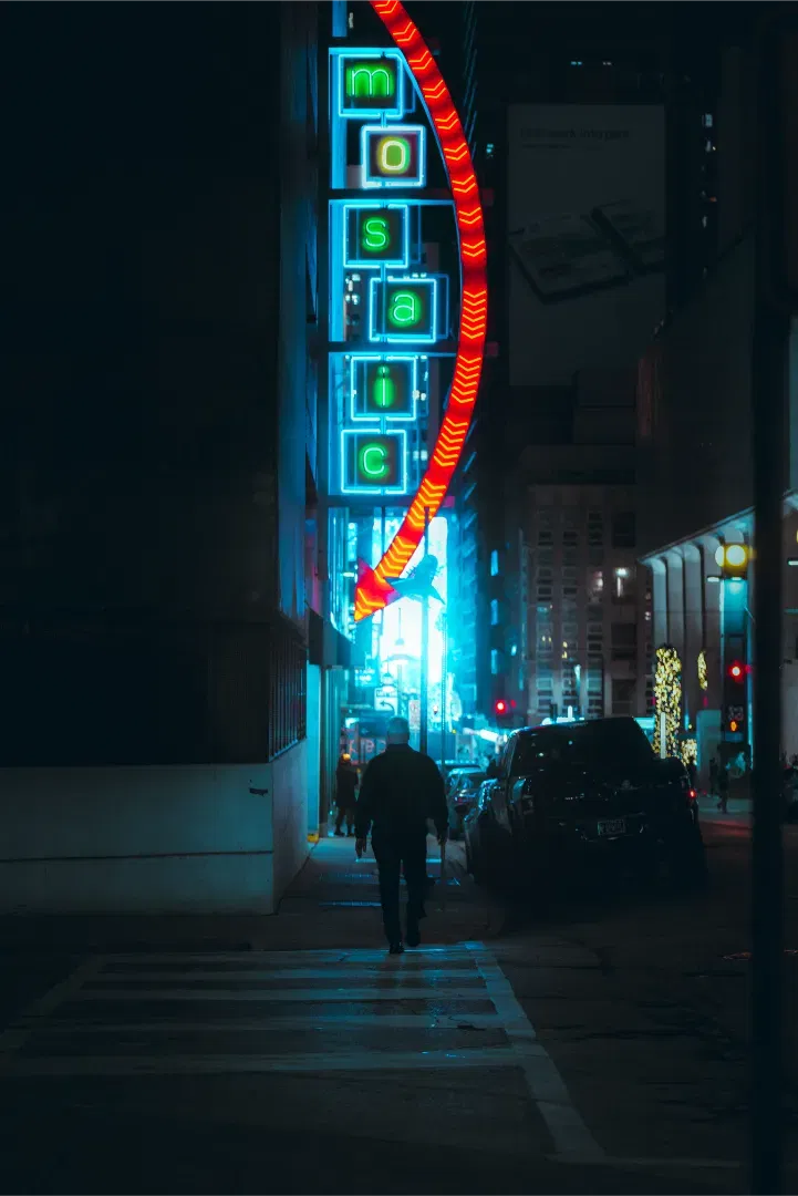 A man walking on a crosswalk at night towards the glowing neon sign of Mosaic building with teal and red tones.
