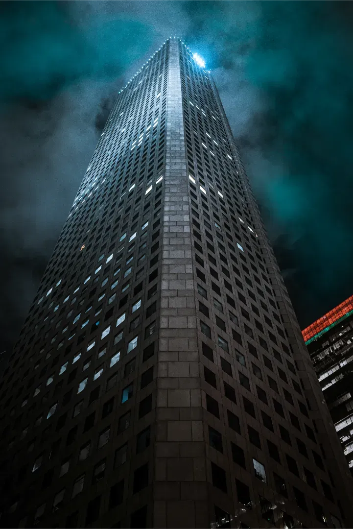Low angle perspective looking up at a towering modern concrete skyscraper against a dark cloudy night sky.