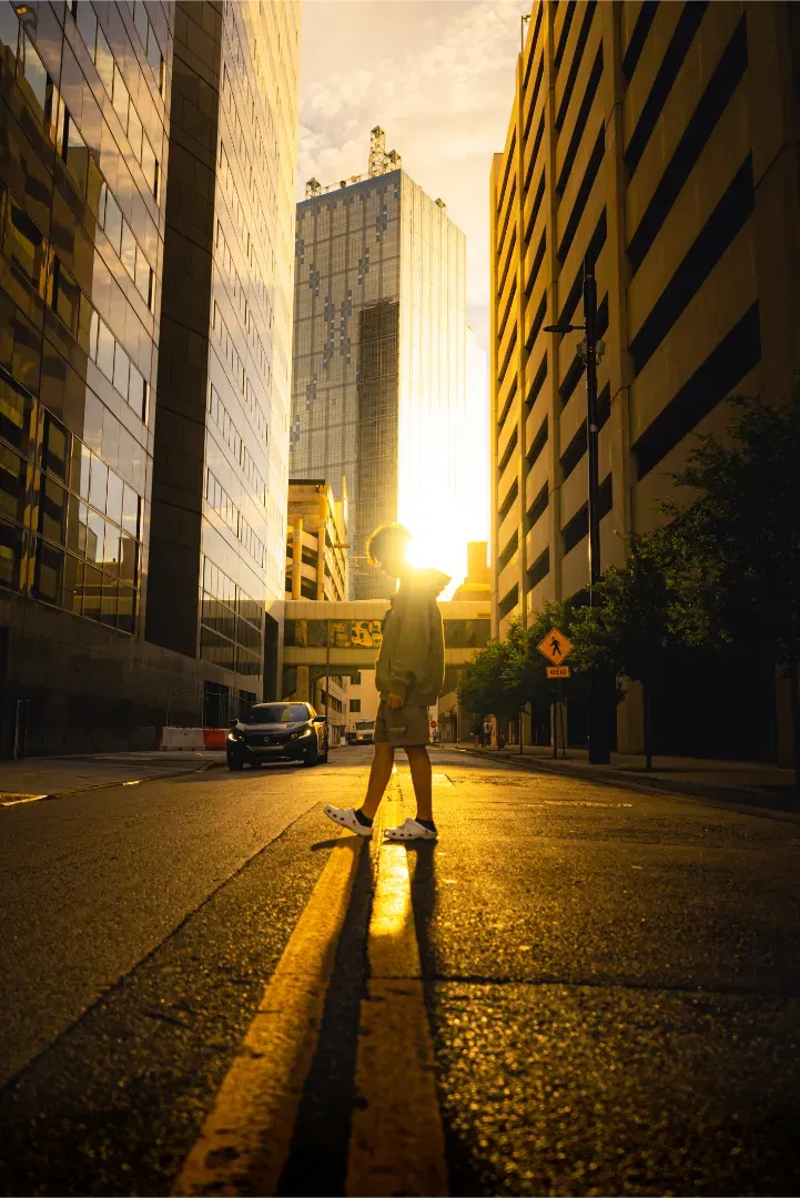 A high-contrast golden hour shot of a pedestrian walking across a city street, with the sun setting behind a building.