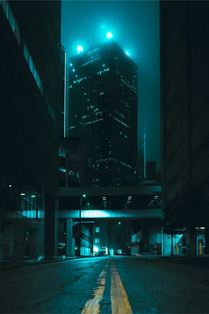 A moody, cyberpunk-style urban street view at night in Dallas, looking down an empty wet road towards a fog-shrouded skyscraper illuminated in cyan.