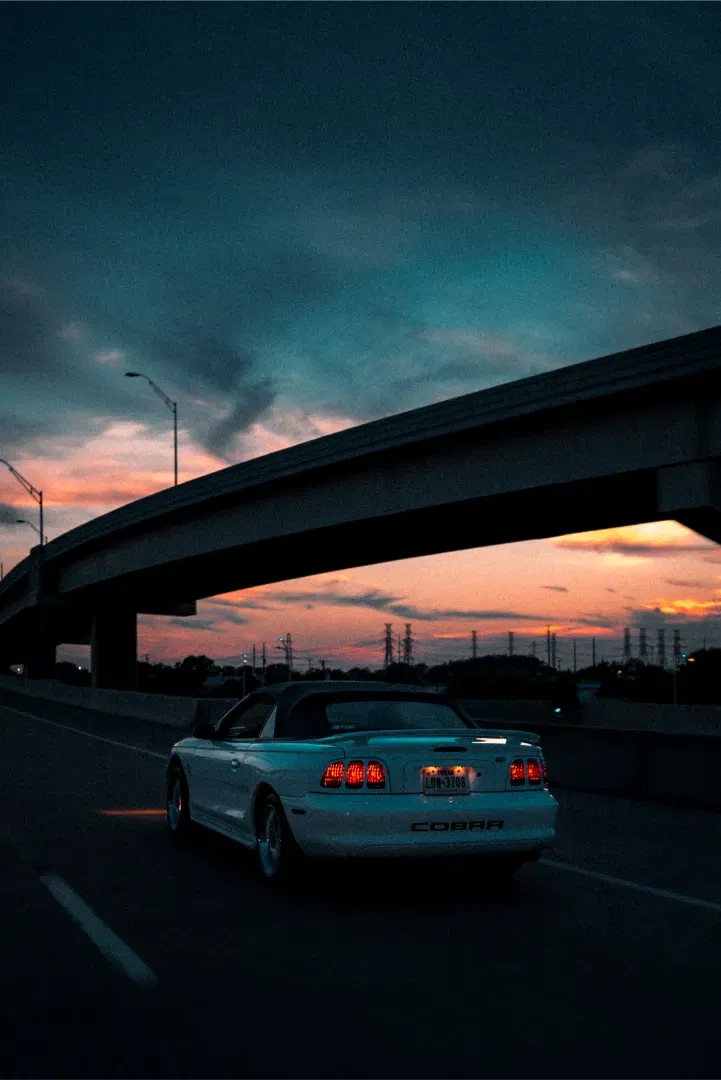 A white Ford Mustang Cobra convertible driving on a highway at sunset, with a concrete overpass above.