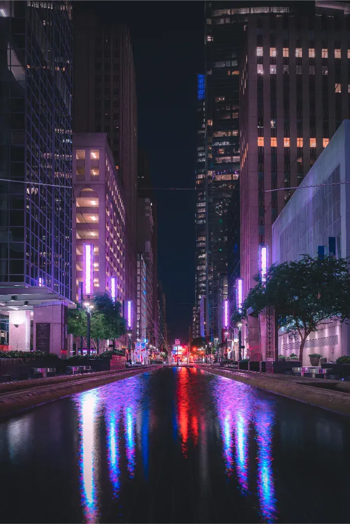 An urban street view at night showing modern high-rises and neon lights reflecting in a pool of water.