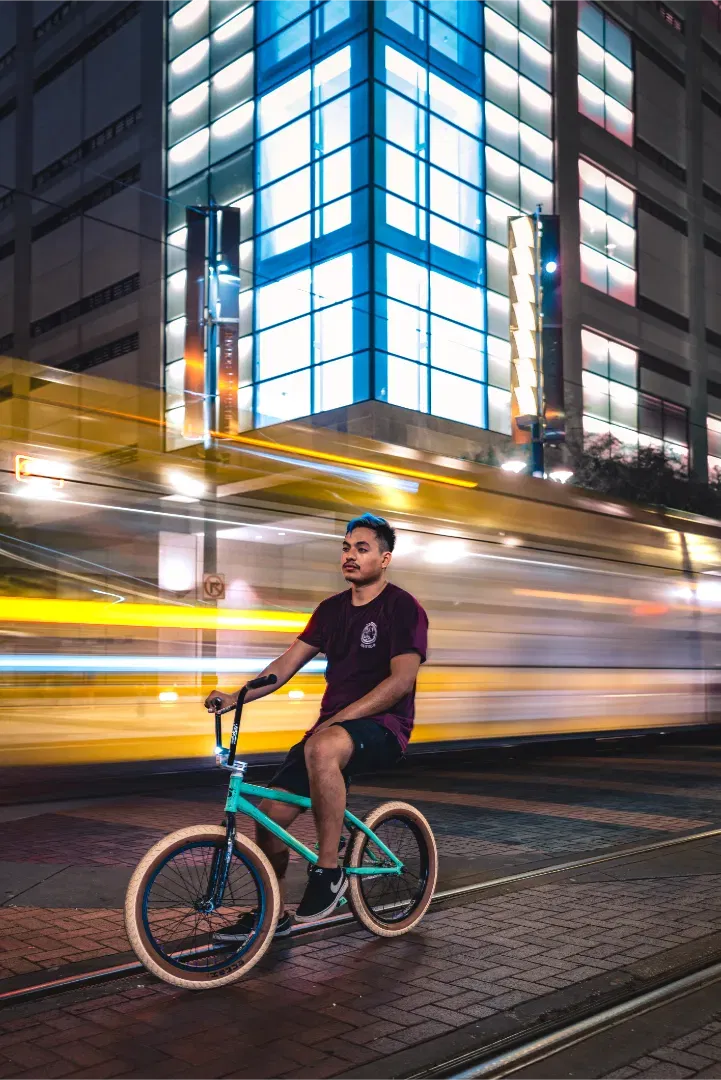 A young man sitting on a teal BMX bike on a city street at night, with the motion blur of a yellow train behind him.