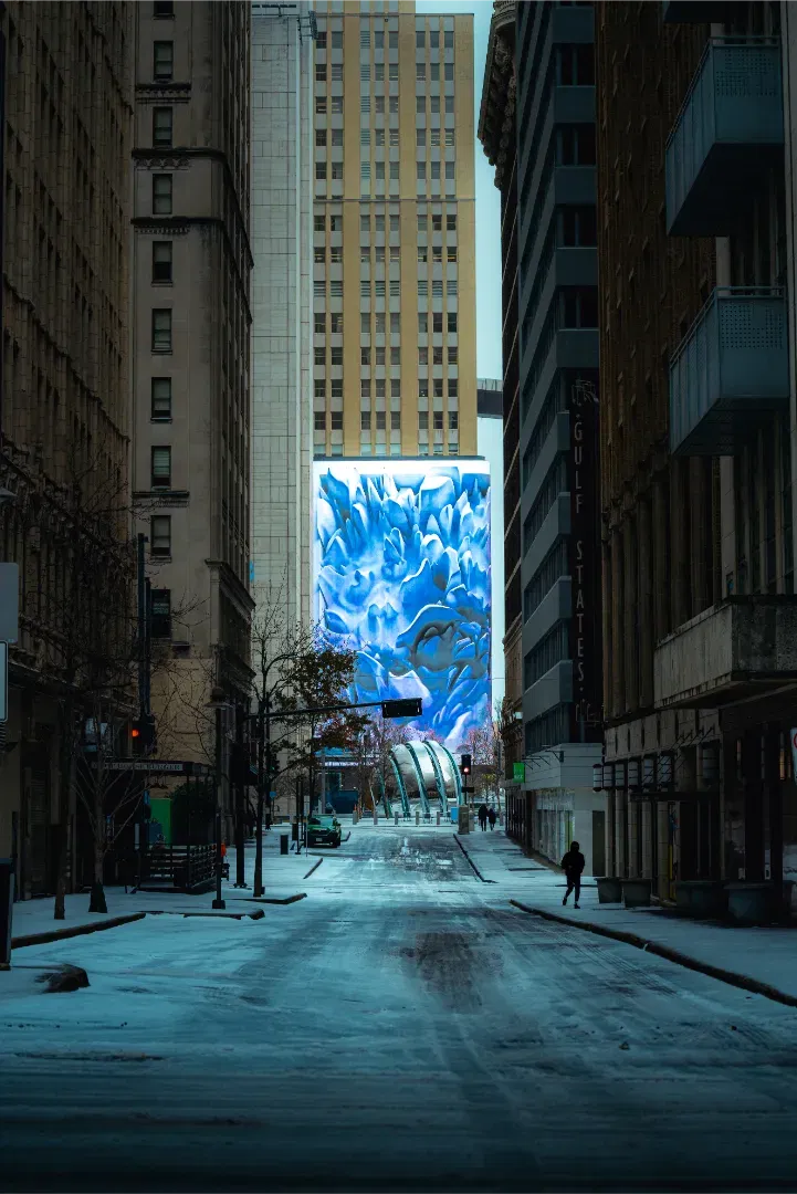 A snow-covered city street looking down an alley flanked by high-rises, featuring a giant blue art installation screen.