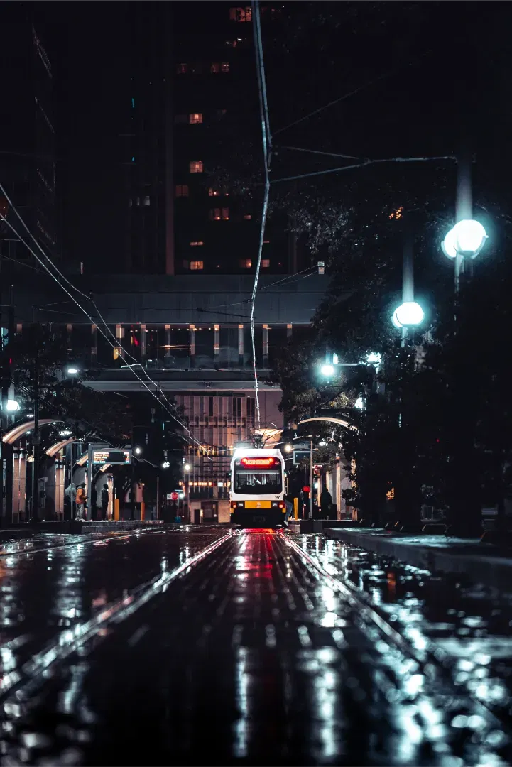 A wet city street at night, reflecting streetlights and a train approaching in the distance.