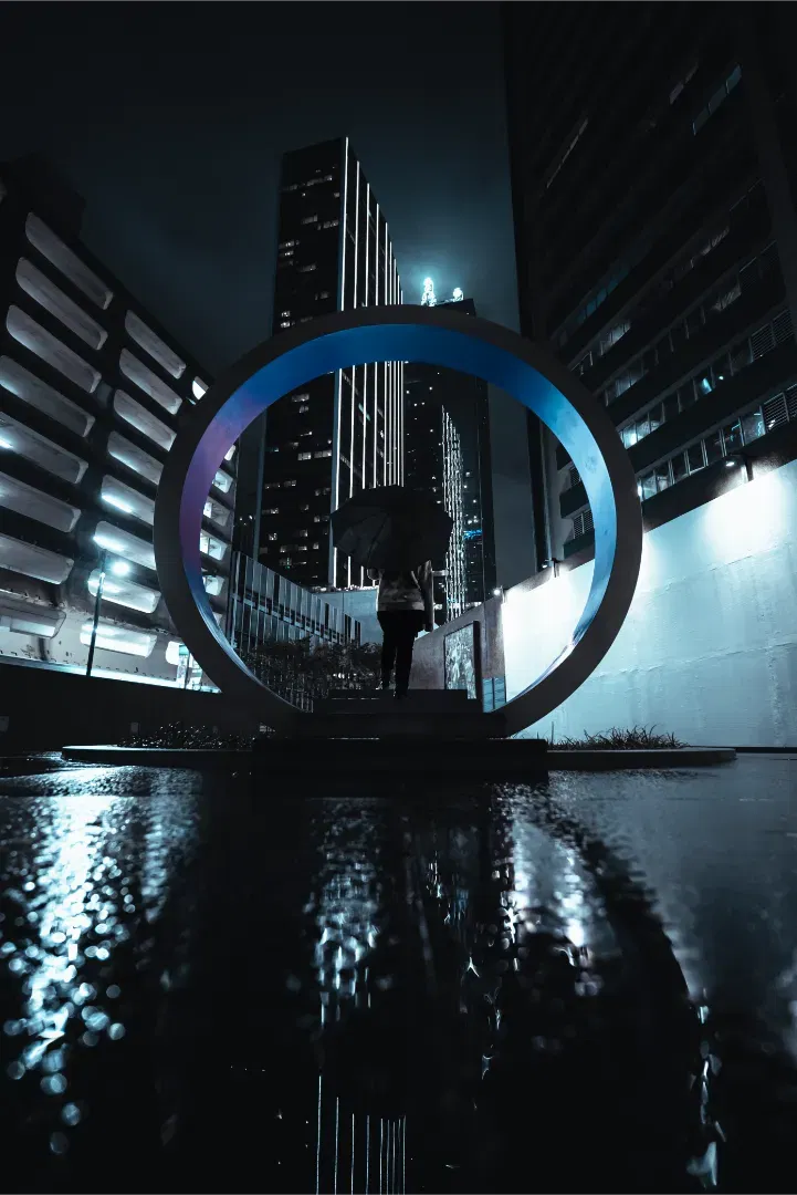 A person with an umbrella standing inside a large circular metallic sculpture on a wet city plaza at night.