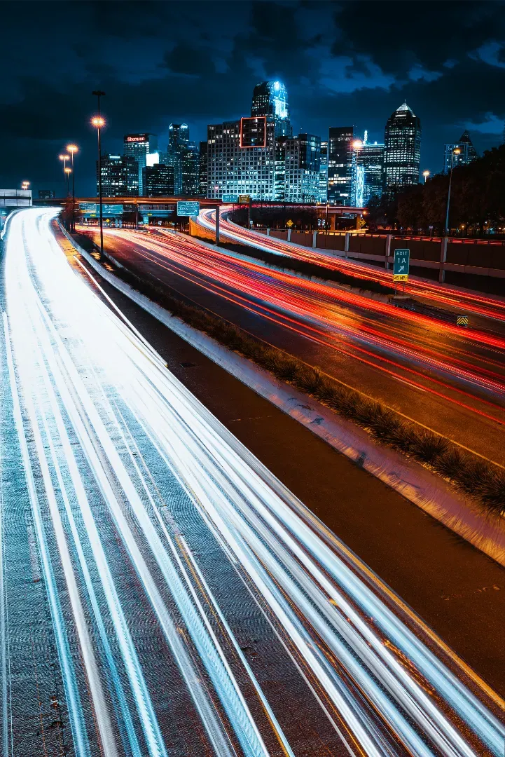 A long exposure night photograph capturing highway traffic light trails leading towards the illuminated Dallas skyline.