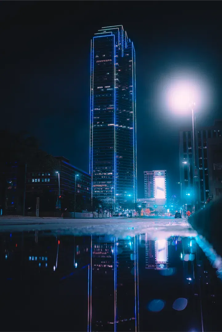Night view of a tall, neon-outlined skyscraper with a bright streetlamp, reflecting in a puddle.