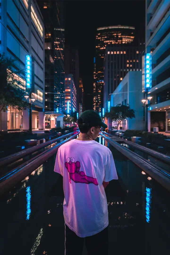 A man in a white t-shirt walking down a wet city street between light-rail tracks reflecting blue neon lights.
