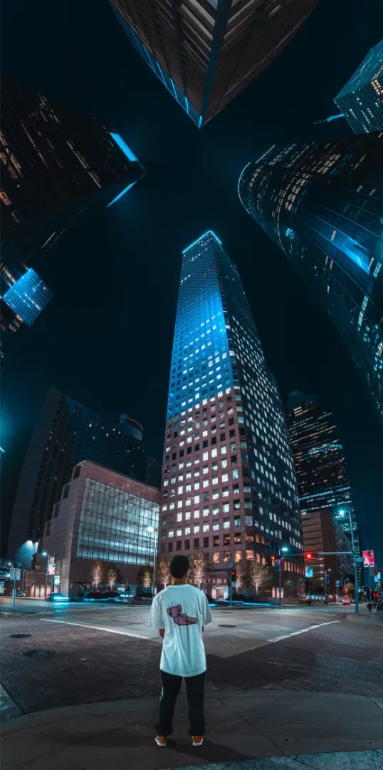 A low angle upward shot of tall skyscrapers at night with a person standing in the crosswalk below looking up.